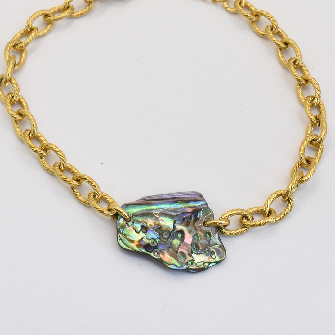 The Salty Dreamers Bracelet | Paua Shell Kowhai features an irregularly shaped pāua shell centerpiece with iridescent green, blue, and purple hues on a gold chain, set against a plain white background.
