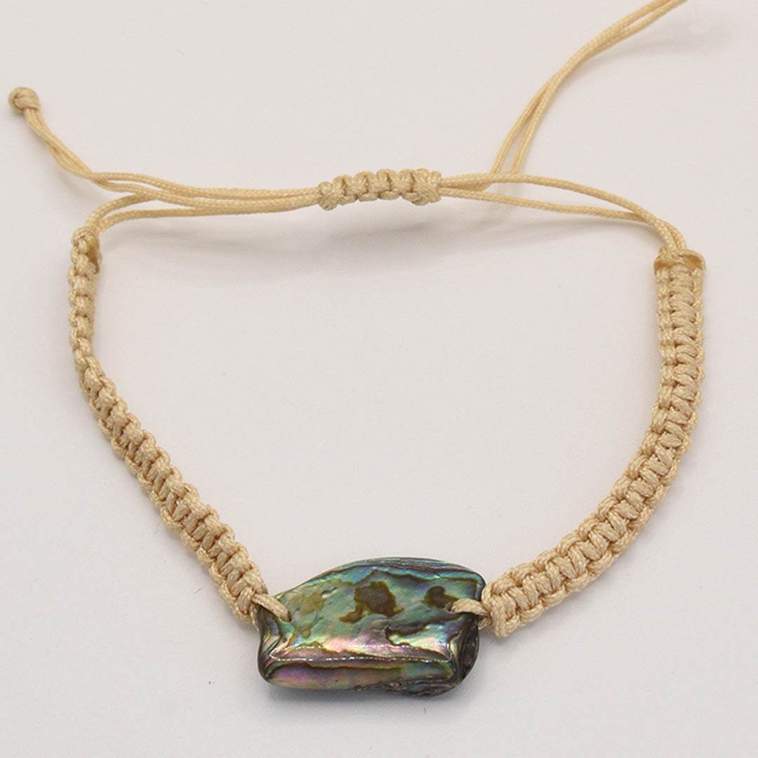 The Bracelet | Paua Shell Rangi by Salty Dreamers is a beige handmade macrame piece with a large iridescent paua shell centerpiece and an adjustable sliding knot, shown against a plain white background.