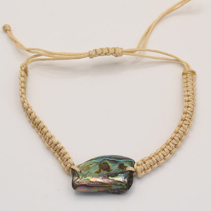 The Bracelet | Paua Shell Rangi by Salty Dreamers is a beige handmade macrame piece with a large iridescent paua shell centerpiece and an adjustable sliding knot, shown against a plain white background.