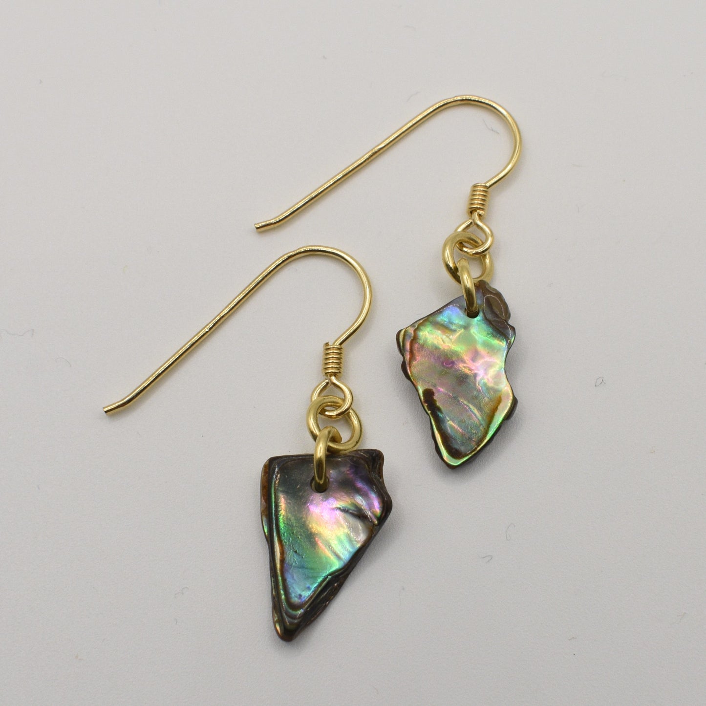 The Salty Dreamers Earrings | Paua Shell Uru feature irregular, iridescent pāua shell pendants on gold hooks, capturing distinctive New Zealand style against a plain white background.