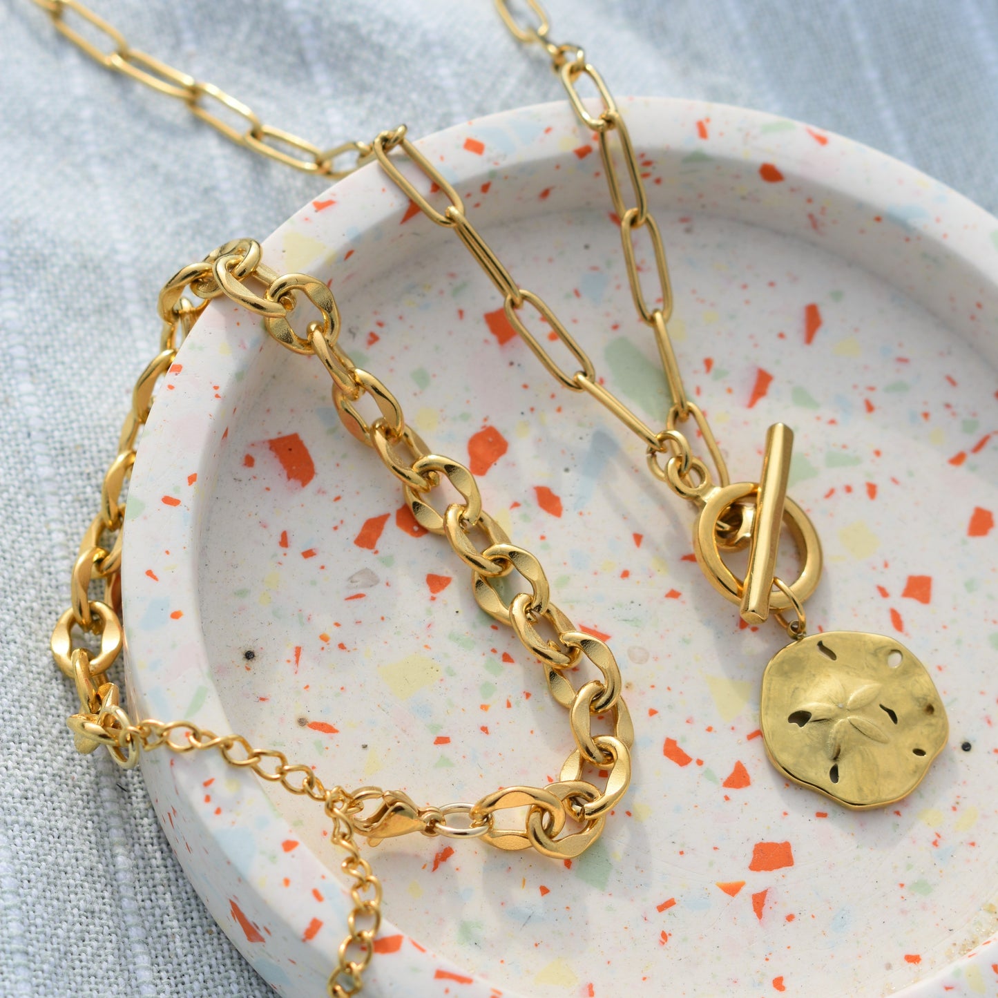 Two gold chain necklaces by Salty Dreamers rest in a round terrazzo dish; one is the Wavy Times ocean necklace with a sand dollar pendant and toggle clasp, while the other features a chunky link design—both beautiful handmade pieces.