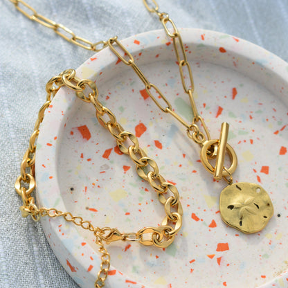 Two gold chain necklaces by Salty Dreamers rest in a round terrazzo dish; one is the Wavy Times ocean necklace with a sand dollar pendant and toggle clasp, while the other features a chunky link design—both beautiful handmade pieces.