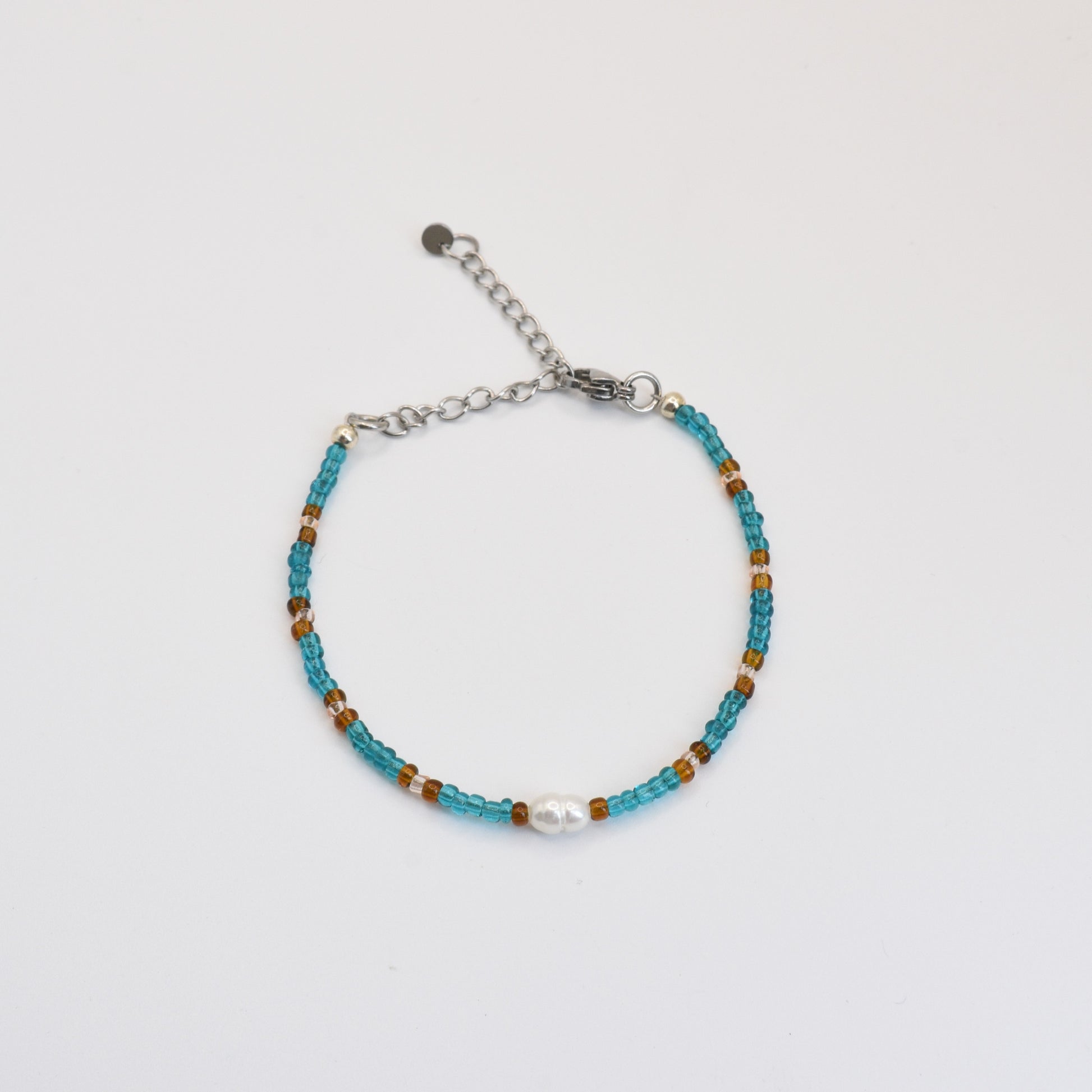 The Bracelet | Ocean Chique by Salty Dreamers features small turquoise and brown beads, a central white freshwater pearl, and an adjustable silver chain clasp, beautifully handmade and displayed on a plain white background.