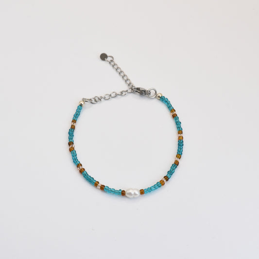 The Bracelet | Ocean Chique by Salty Dreamers features small turquoise and brown beads, a central white freshwater pearl, and an adjustable silver chain clasp, beautifully handmade and displayed on a plain white background.