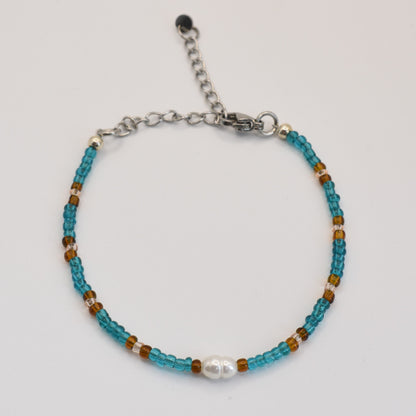 The Bracelet | Ocean Chique by Salty Dreamers features handmade turquoise and brown beads, a central white pearl-like bead, and an adjustable silver clasp—ideal for surfer chic style. Shown against a white background.