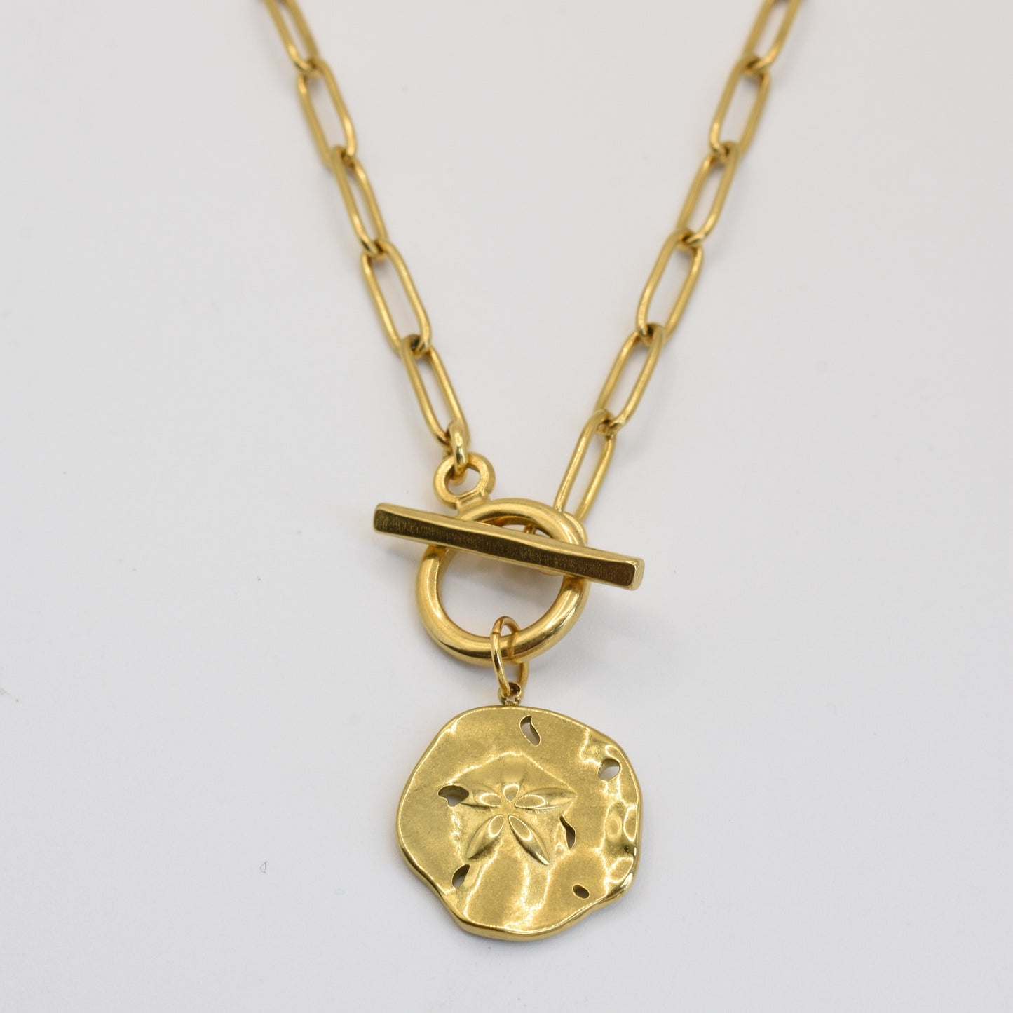 The Wavy Times necklace by Salty Dreamers features a gold, textured round pendant with a small flower design and elongated link chain with a toggle clasp—an elegant handmade accessory for any occasion.