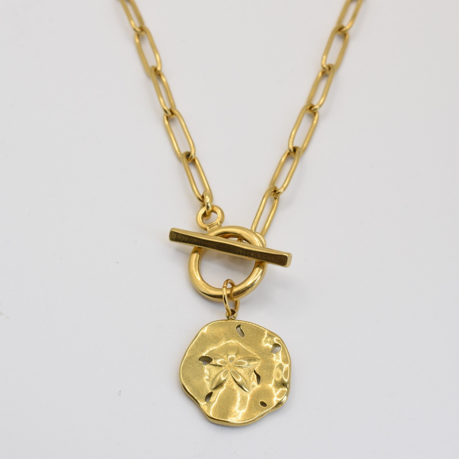 The Wavy Times necklace by Salty Dreamers features a gold, textured round pendant with a small flower design and elongated link chain with a toggle clasp—an elegant handmade accessory for any occasion.