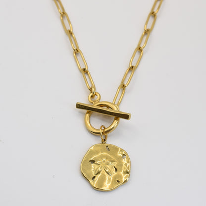 The Wavy Times necklace by Salty Dreamers features a gold, textured round pendant with a small flower design and elongated link chain with a toggle clasp—an elegant handmade accessory for any occasion.