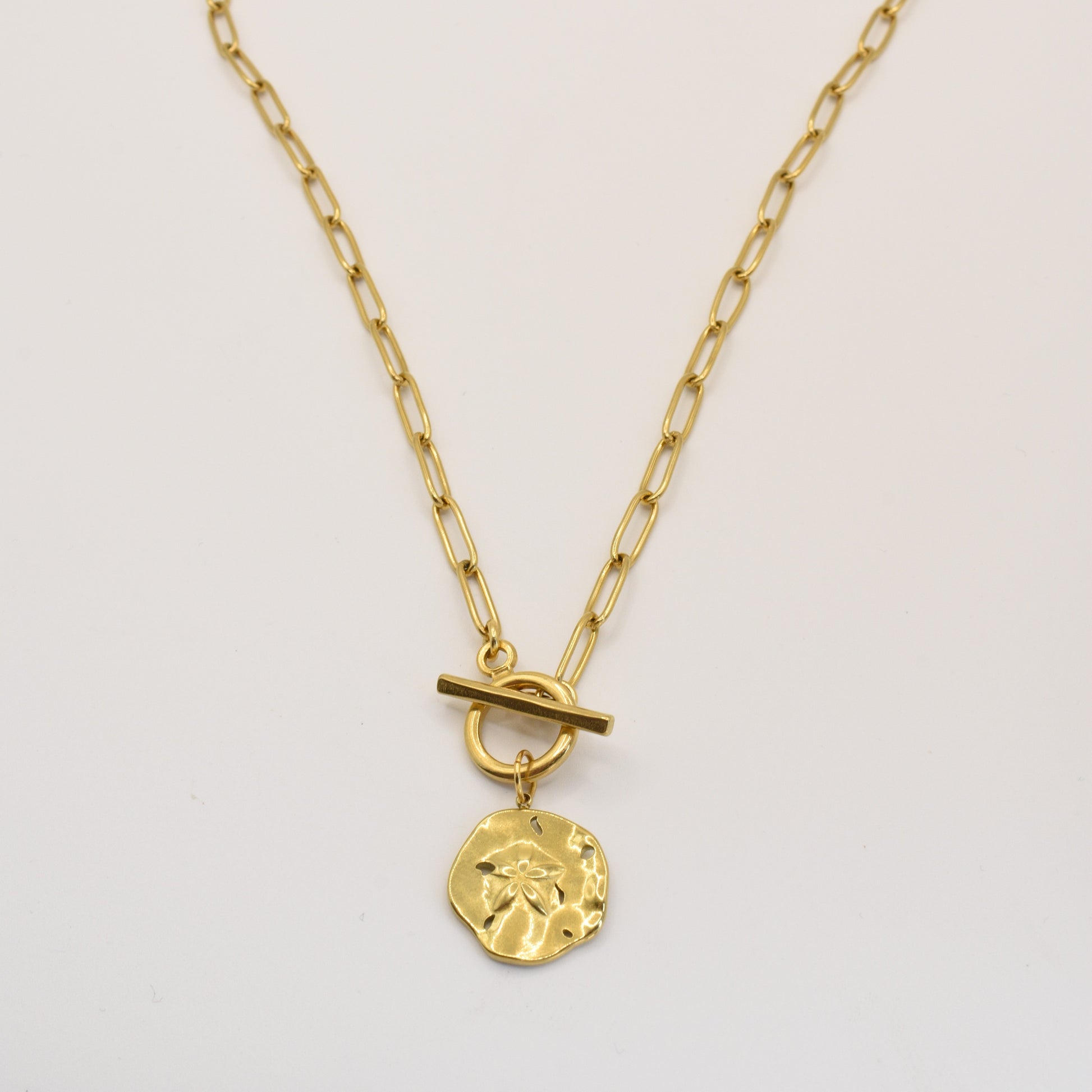 The Wavy Times Necklace by Salty Dreamers features elongated gold links and a round, hammered starburst pendant with a central toggle clasp. This handmade jewelry piece is photographed on a plain white background.