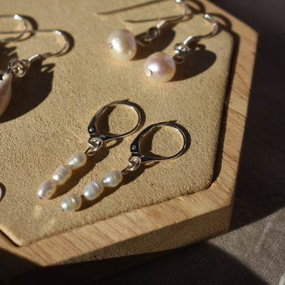 Two pairs of Salty Dreamers Pretty Pearly Earrings are displayed on a tan hexagonal tray. One pair has three freshwater pearls on 925 silver hoops, while the other features single round pearls on hooks, softly lit by sunlight.