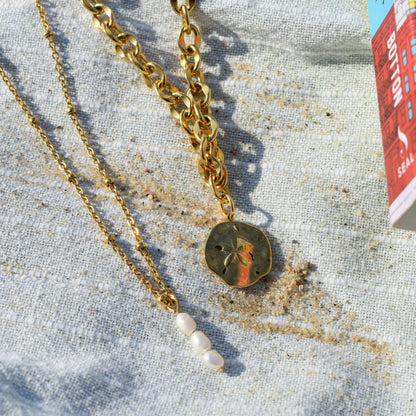 Two gold necklaces from Salty Dreamers rest on light textured fabric: one is the Ocean Dollar Gold necklace with a sand dollar pendant, and the other has a delicate chain with three small white pearls. Sand and part of a colorful book are nearby.