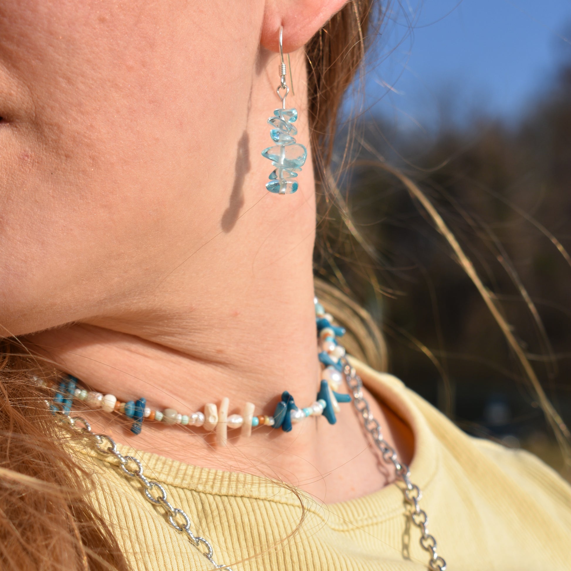 A close-up of a person wearing Salty Dreamers Necklace | Ocean Bamboo Blue, featuring blue stones and pearls, styled with a yellow shirt and sunlit hair.