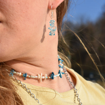 A close-up of a person wearing Salty Dreamers Necklace | Ocean Bamboo Blue, featuring blue stones and pearls, styled with a yellow shirt and sunlit hair.