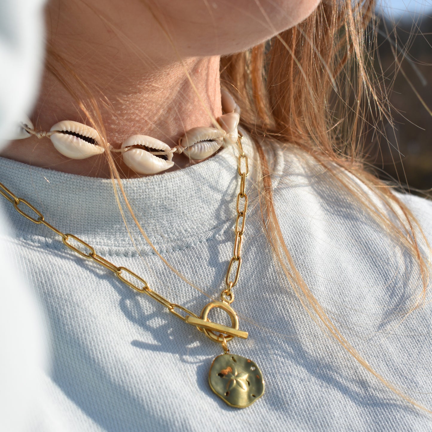 Close-up of a person wearing a white shirt and the Salty Dreamers Necklace | Wavy Times, featuring a gold chain with a round, textured pendant. A shell choker completes the look, as light brown hair frames a blurred outdoor background.