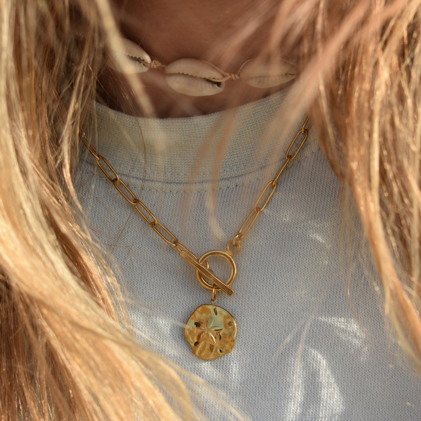 Close-up of a person wearing the Salty Dreamers Necklace | Wavy Times, featuring a gold chain with a round, textured pendant and a white shell choker. Their wavy blonde hair partially covers the handmade jewelry worn over a light top.