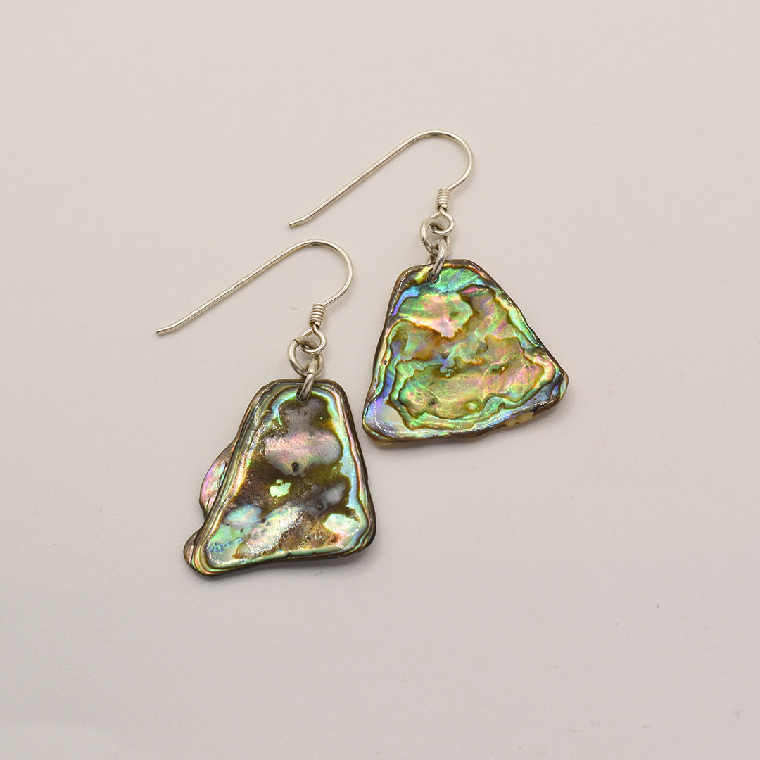 Salty Dreamers Earrings | Paua Shell Kea: Iridescent, irregularly shaped pāua shell earrings with silver hooks, displayed on a light background—a unique touch of New Zealand jewelry.