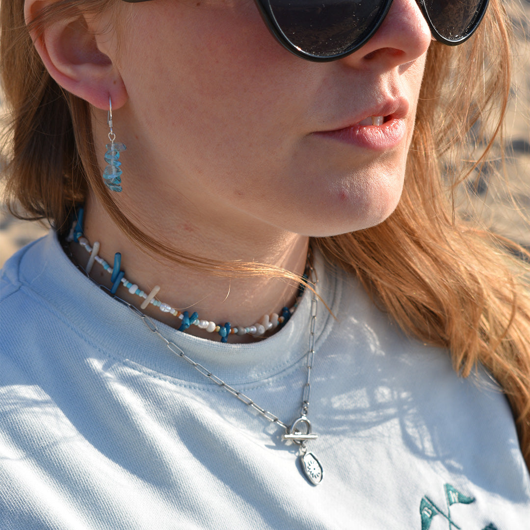 A woman in sunglasses and a light blue shirt wears Salty Dreamers Glassy Spring 925 silver earrings, layered choker and necklace, with sunlight highlighting her face and hair.