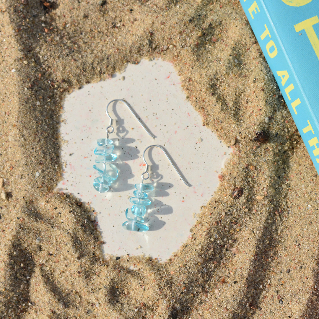 The Glassy Spring earrings by Salty Dreamers, featuring a light blue spiral design, are displayed on sand beside a blue and yellow book, resting in a smooth cleared space surrounded by grains of sand.