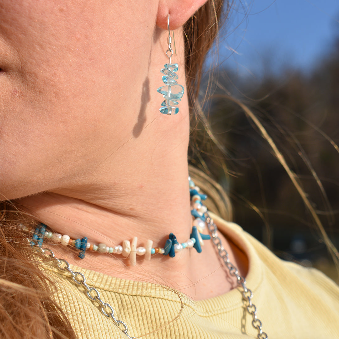 A close-up of a person wearing Salty Dreamers Glassy Spring bear-shaped glass earrings, paired with layered necklaces featuring blue and white beads, shells, and silver chains against a blurred outdoor background.