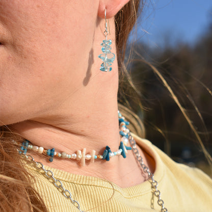 A close-up of a person wearing Salty Dreamers Glassy Spring bear-shaped glass earrings, paired with layered necklaces featuring blue and white beads, shells, and silver chains against a blurred outdoor background.