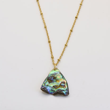 The Salty Dreamers Paua Shell Tai Necklace features a handmade pendant with an iridescent, irregular paua shell in green, blue, and purple hues, displayed on a gold chain against a plain white background.