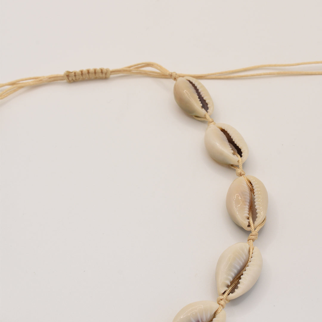 A close-up of the Salty Dreamers Necklace | Cowrie Choker shows a beige braided cord adorned with cream cowrie shells, arranged in a row on a white background.