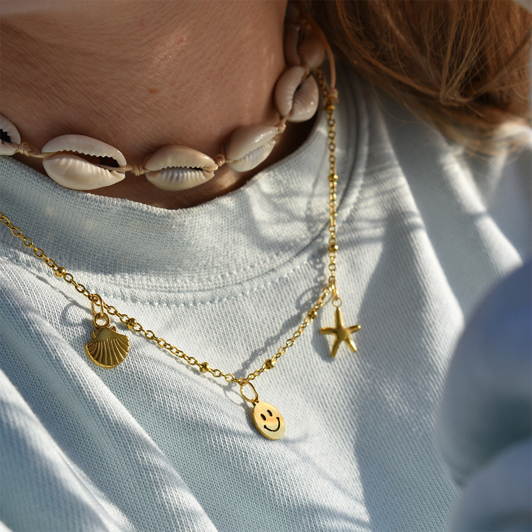 A person wearing a light blue shirt and the Salty Dreamers Necklace | Cowrie Choker, layered with two gold chain necklaces featuring seashell, starfish, and smiley face beach charms.
