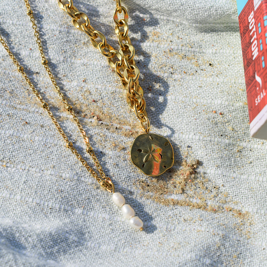 Two gold necklaces from Salty Dreamers rest on sandy fabric—a chunky chain with a round pendant and the Necklace | Cutie Pearlies, a handmade pearl necklace with three small pearls. A red and blue box peeks in from the upper right.