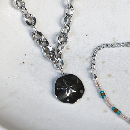 The Salty Dreamers Ocean Dollar Silver necklace with a chunky stainless steel chain and sand dollar pendant rests beside a delicate turquoise-accented beaded bracelet on a white ceramic dish.