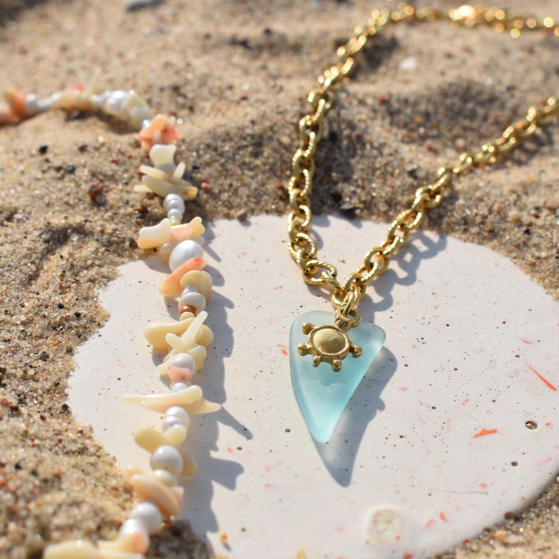 The Salty Dreamers Turtle Sea Glass Necklace rests on sandy ground beside a shell and bead bracelet, both displayed on a white, round stone.