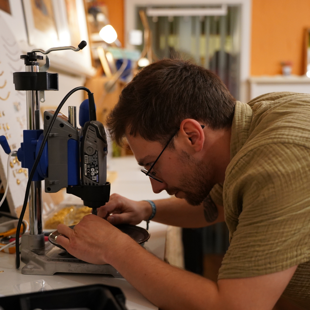 A man with glasses and a short beard uses a drill press on a small object in the well-lit Salty Dreamers Workshop Meeresschmuck in Kiel, focusing intently as tools and materials are arranged on the table around him.